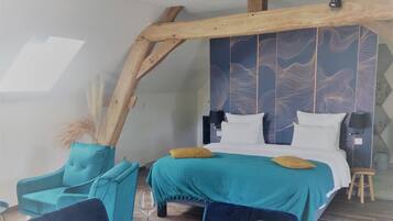 Ein gemütliches Schlafzimmer mit schrägverdeckter Decke, einem Bett mit türkisfarbenem Bettzeug, einem passenden Sessel und einem kleinen Hocker.
