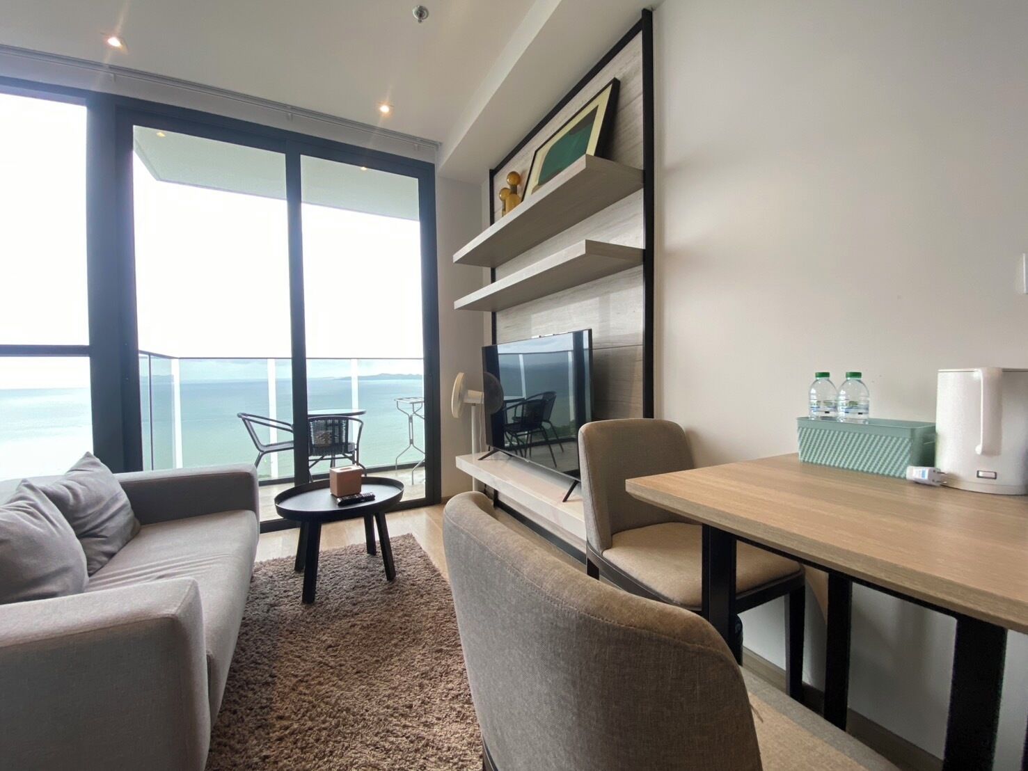 Ein modernes Wohnzimmer mit Sofa, Sessel, Couchtisch und einem Essbereich mit Tisch und Stühlen. Ein Fernseher steht auf einem Schrank, es gibt ein Bücherregal und durch große Fenster blickt man auf den Ozean.