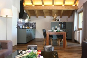 Eine moderne Küche mit Holzbalken, ein Essbereich mit Tisch für vier Personen und ein Wohnzimmer mit Sofa und Lampe.
