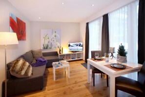 Ein modernes Wohnzimmer mit einer Couch, einem Couchtisch, einem Esstisch für zwei Personen und einem Fernseher im Hintergrund.