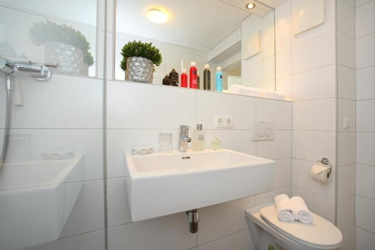 A modern bathroom with a white sink, a toilet, and a shower. There are toiletries on the sink and a potted plant on the wall.