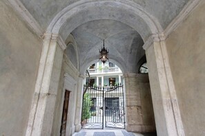 Un pasillo arqueado con un candelabro, una entrada con reja y vista a un edificio con columnas y ventanas.