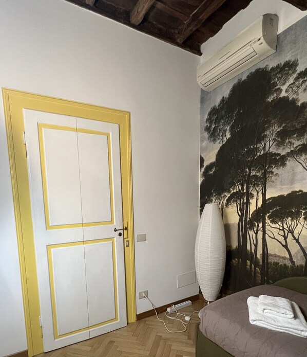 Una habitación con puerta doble, unidad de aire acondicionado, un cuadro con árboles, una lámpara y una cama con una toalla.