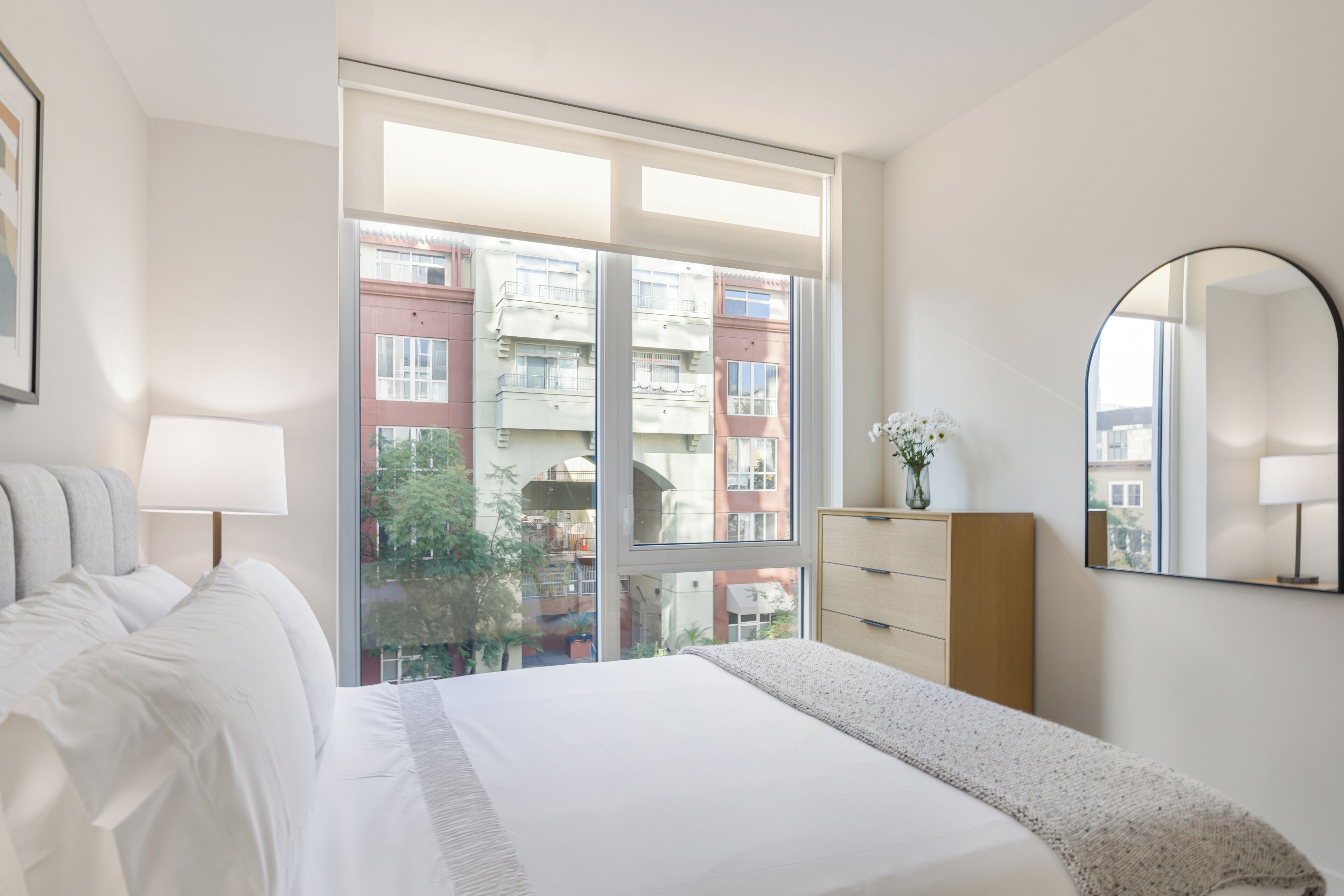 A hotel room with a large window, a bed, a dresser, and a mirror.