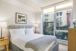 A modern bedroom with a bed, bedside tables, a window with a view of buildings, and a framed abstract painting. - Modern 1BR + Rooftop Pool + Gym | Near the Bay | AvantStay Little Italy (San Diego)