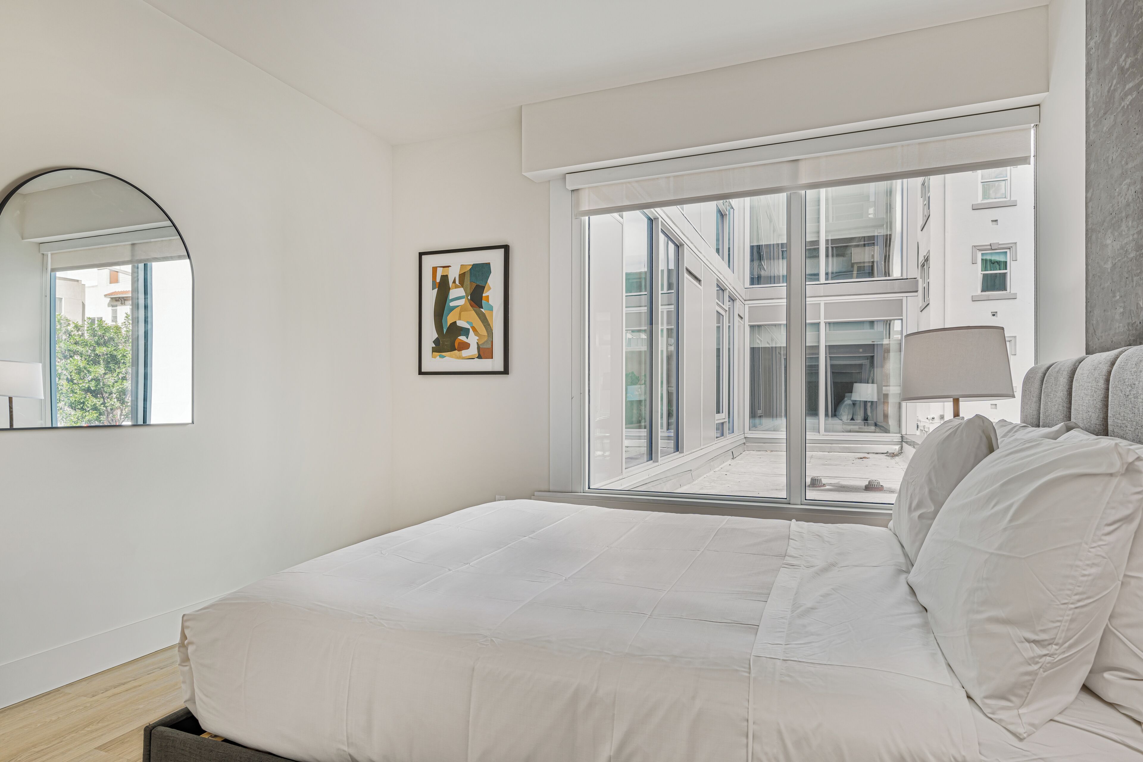 Un dormitorio moderno con una cama grande, un ventanal con vistas a edificios, un cuadro abstracto enmarcado y un espejo en la pared.