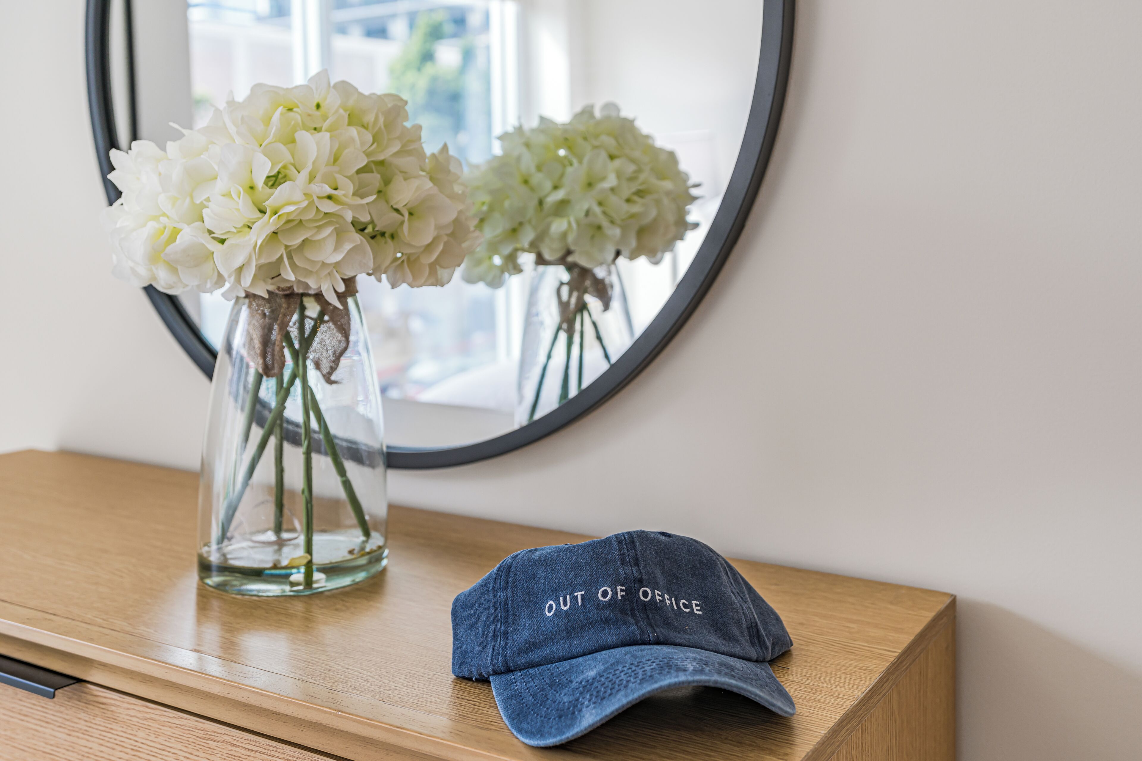 Una mesa de madera con un jarrón de cristal transparente que contiene flores blancas y una gorra vaquera con la frase 'OUT OF OFFICE'.