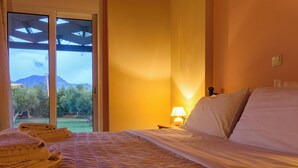 Ein Hotelzimmer mit Bett, Nachttisch, Lampe und Blick auf einen Garten und Berge durch eine Glastür.