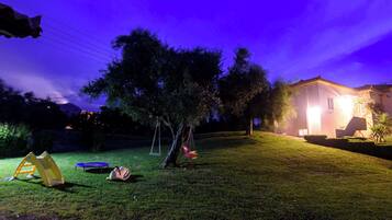 Un'area verdeggiante con un albero, altalene e una casa sullo sfondo.