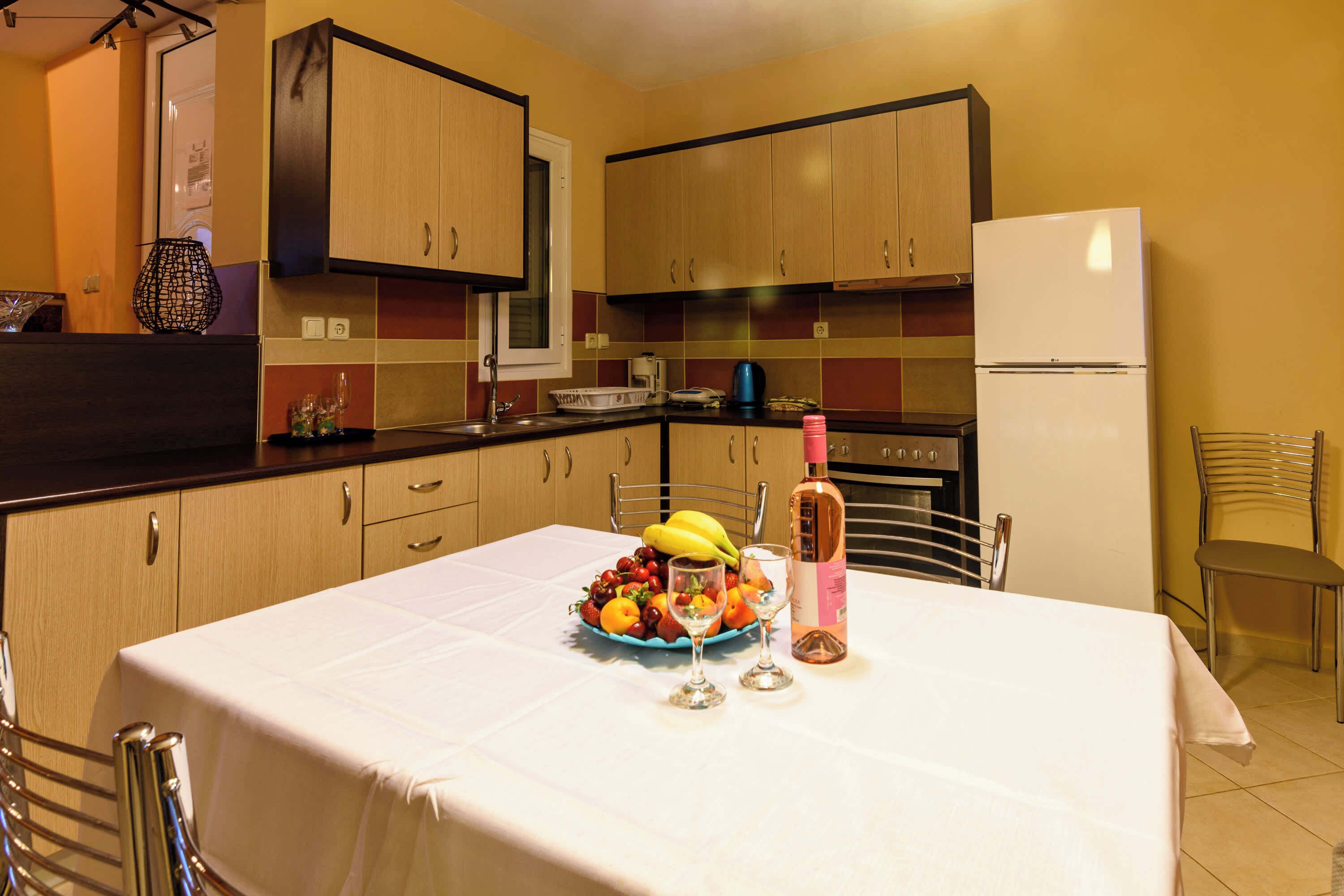Een keuken met houten kastjes, een witte koelkast en een eettafel gedekt met een fruitschaal, wijnkaraffen en een fles.