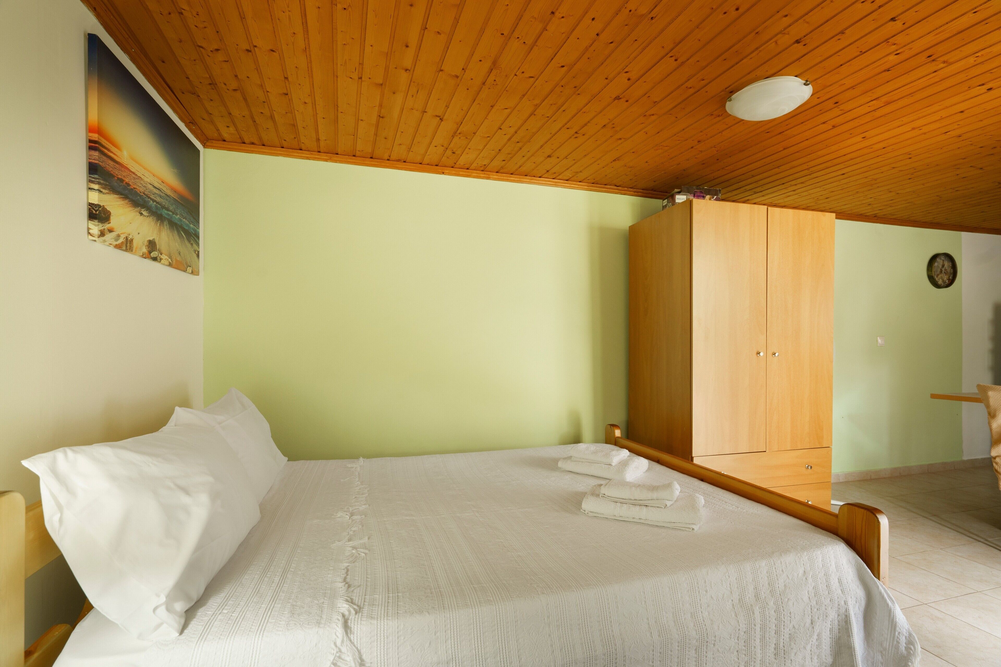 A bedroom with a wooden ceiling, a bed with white linens, a wooden wardrobe, and a painting on the wall.