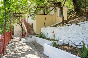 A stone pathway leading to a building with a staircase, surrounded by greenery and a white wall.