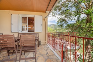 A balcony with a wooden table and chairs, a red railing, and a view of greenery and hills.