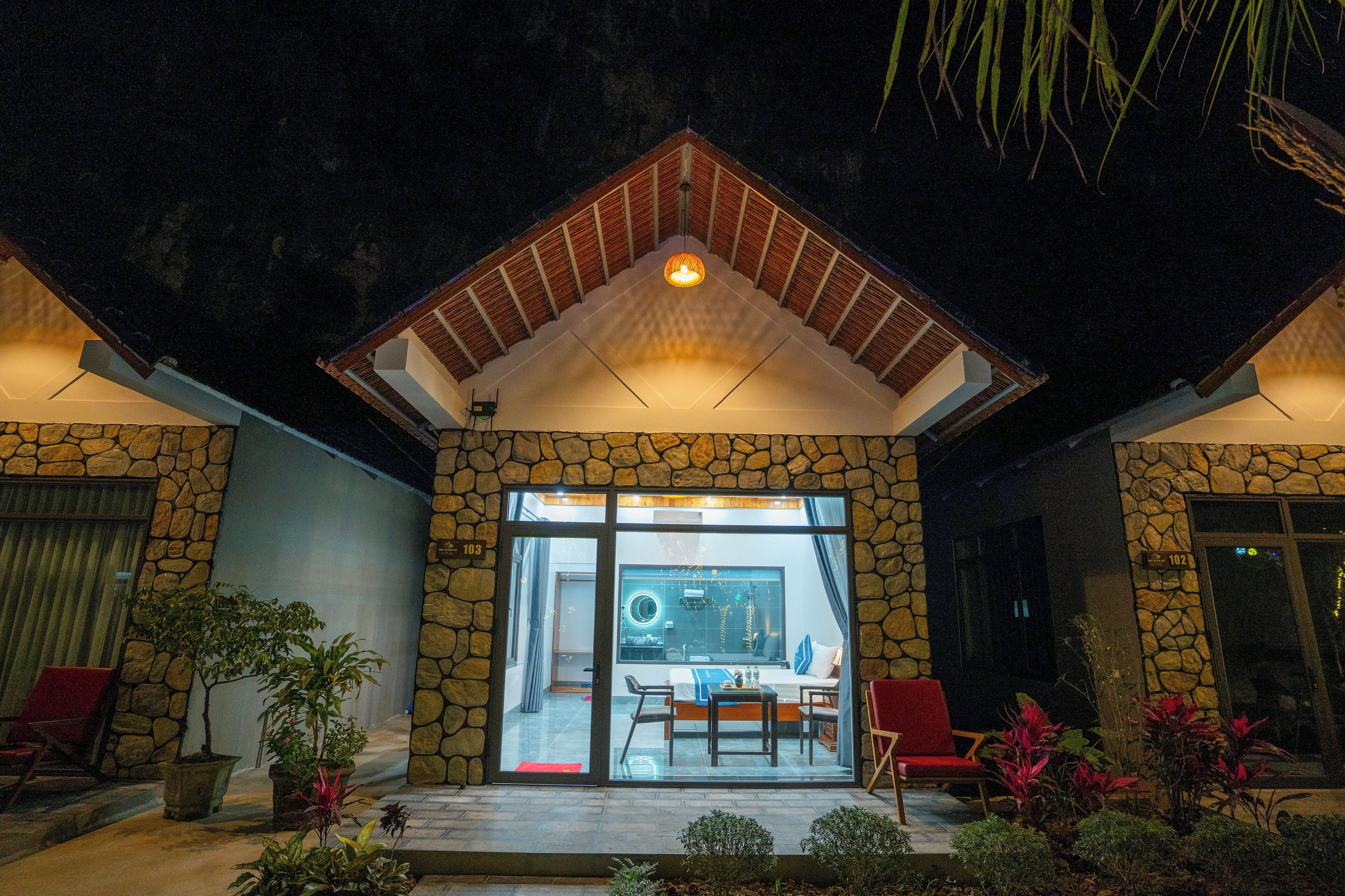 A stone building with a glass door entrance, a small patio area with chairs, and a bed visible inside.