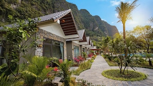 A row of buildings with stone facades and tiled roofs, surrounded by lush greenery and a mountainous backdrop.