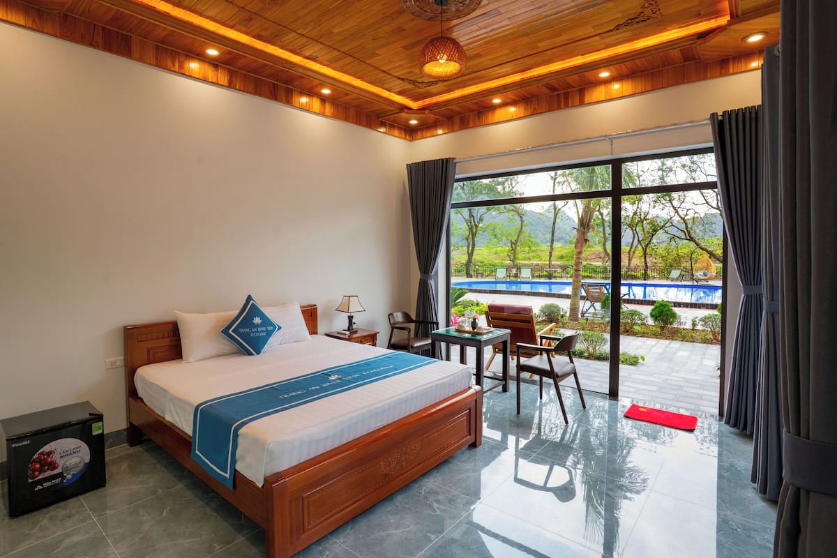 A hotel room with a wooden ceiling, a large bed with a blue bedspread, a view of a pool and greenery outside, and a small table with chairs.