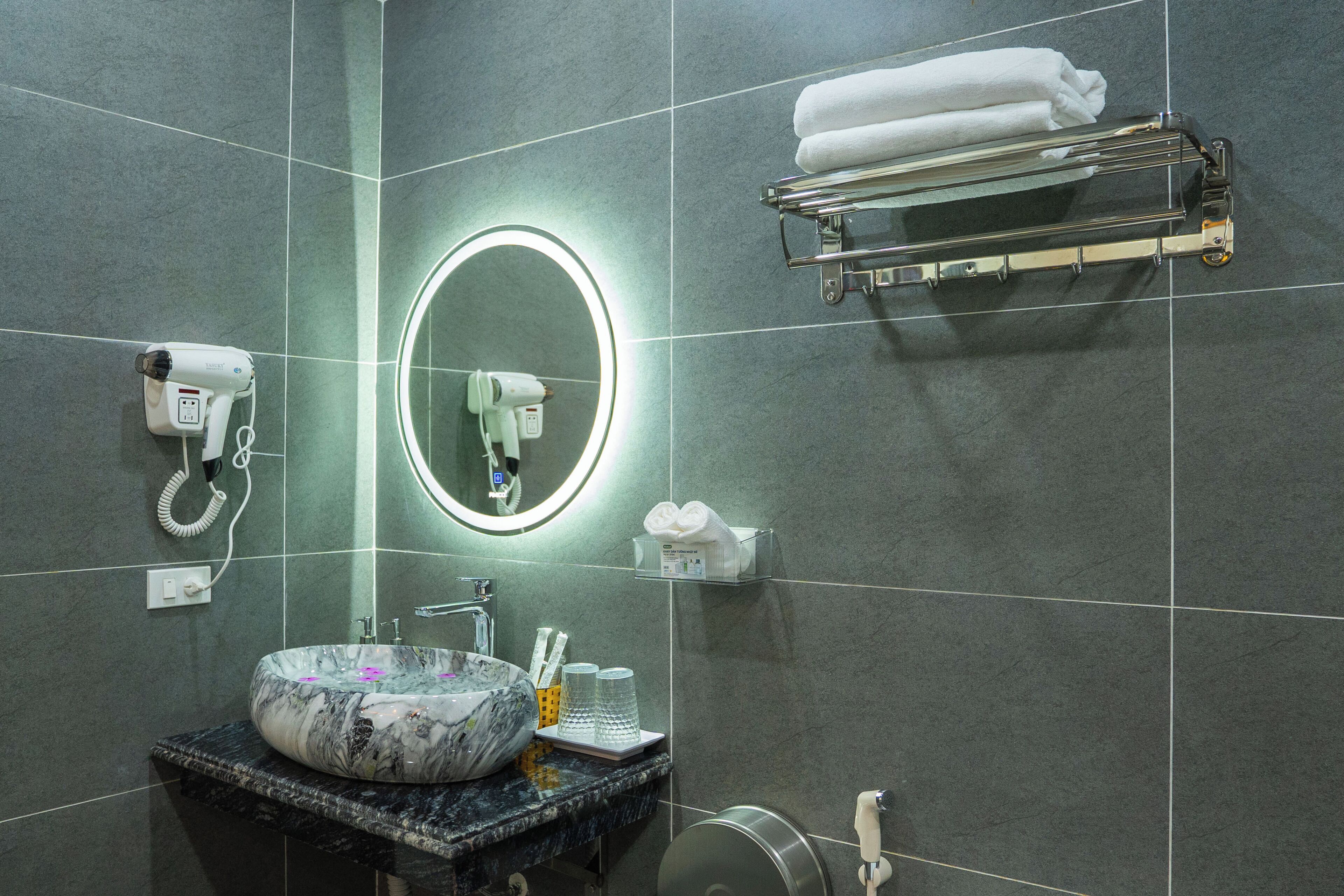 Une salle de bain avec un lavabo en marbre, un miroir rond avec un éclairage intégré, un sèche-cheveux, un porte-serviettes et un distributeur de savon.