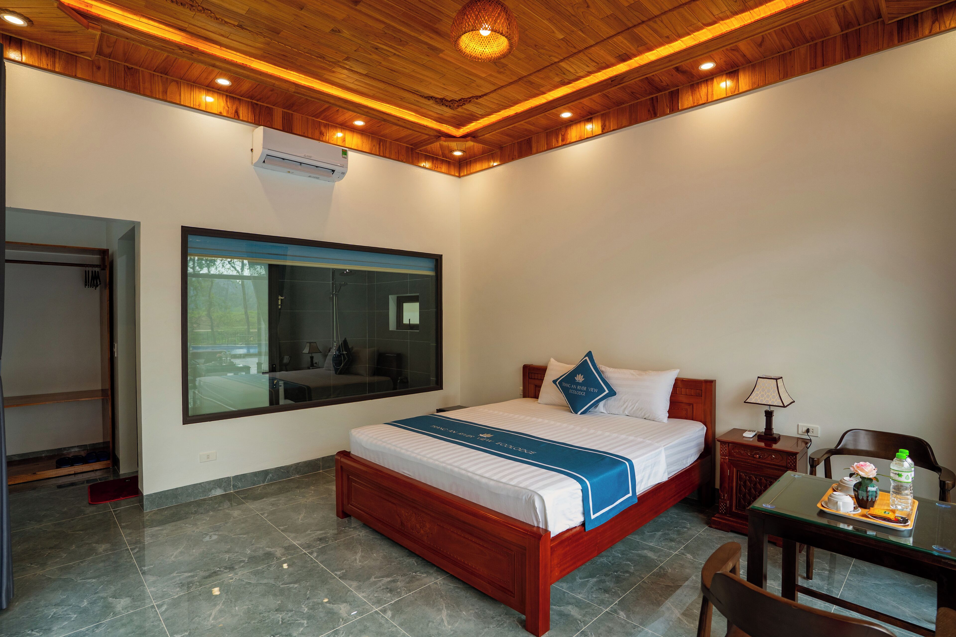 A hotel room with a wooden ceiling, a large window, a bed with a blue bedspread, a nightstand with a lamp, and a table with a tray.
