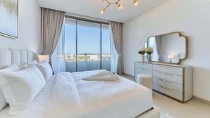Quarto moderno com uma cama grande, cômoda, espelho e vista da cidade através de amplas janelas.