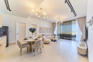 A modern living room with a dining area, a sofa, a coffee table, and a view of the outdoors. - AL Zorah 1 Bedroom Sea and Community View (Ajman)