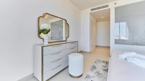 Quarto moderno com penteadeira branca, um banquinho redondo, cama com roupa de cama branca e um espelho grande.