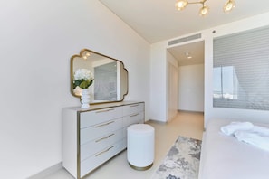 Een moderne slaapkamer met een witte dressoir, een rond krukje, een bed met witte lakens en een grote spiegel.