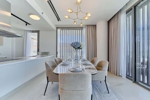 Een moderne eetruimte met een ronde tafel voor vier personen, omringd door gestoffeerde stoelen. Een kroonluchter hangt aan het plafond, en er is een groot raam met gordijnen zichtbaar.