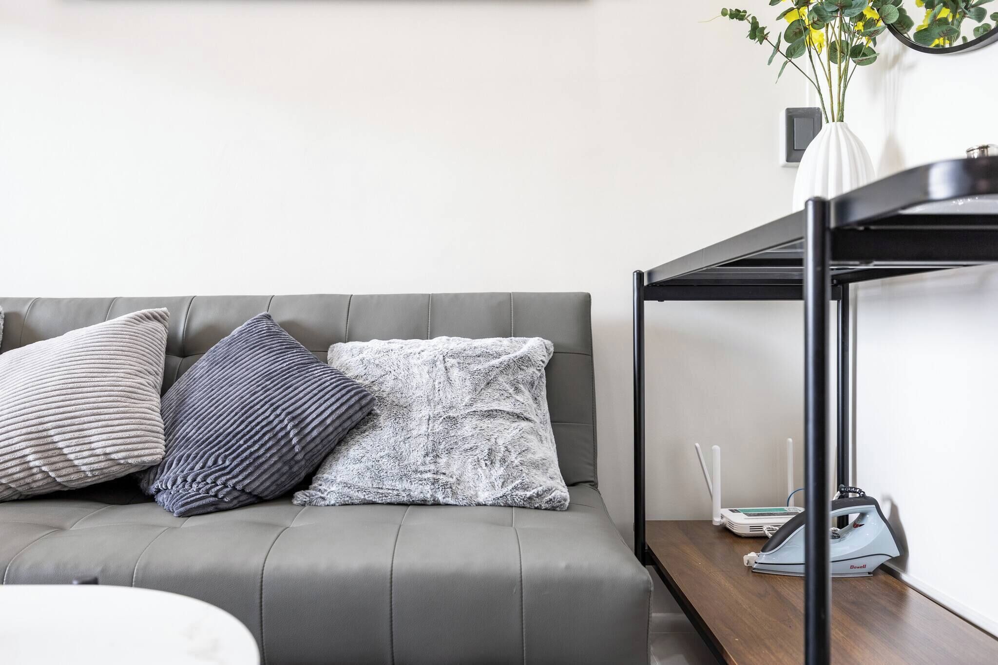 Un sofá gris con tres almohadas, una mesa de metal negro con un jarrón blanco y un dispositivo, y un piso de madera.