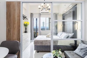 Ein modernes Schlafzimmer mit einem großen Bett, einer Couch, einem kleinen Tisch und Blick auf die Stadtlandschaft durch große Fenster.