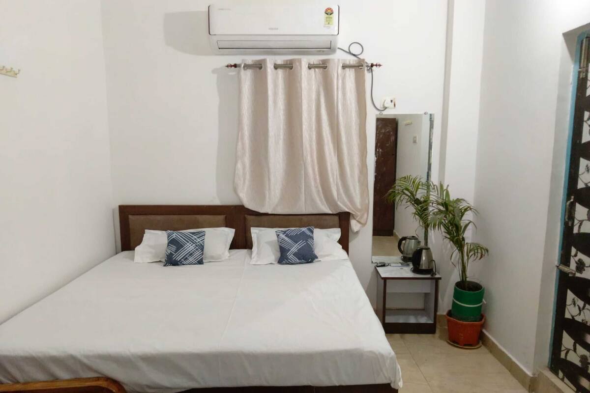 A single bed with a wooden headboard, white bedding, and two patterned pillows. There is a small bedside table with a kettle and a potted plant. An air conditioning unit is mounted on the wall above the bed.