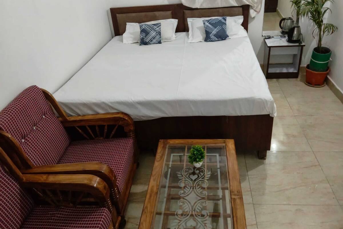 A bedroom with a bed, a wooden sofa, a glass coffee table with a plant, and a small bedside table with a kettle and a plant.