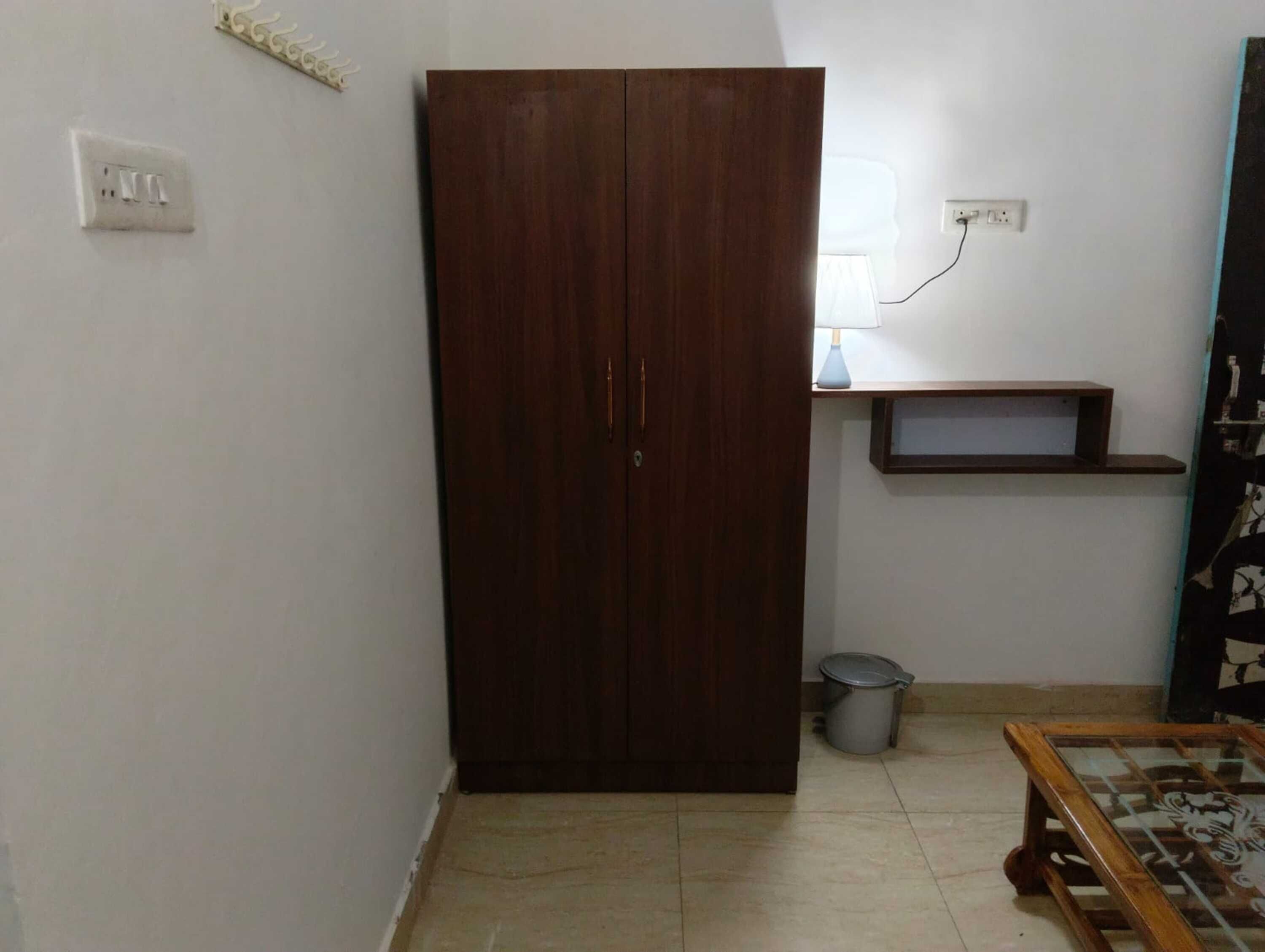 Una habitación con armario, una estantería pequeña, lámpara, cubo de basura y mesa.