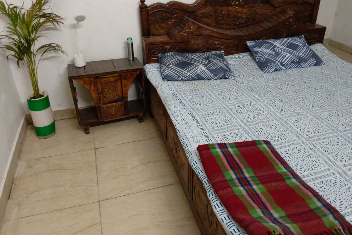 A bed with a patterned bedspread, two pillows, a bedside table with a lamp, a potted plant, and a small wooden nightstand.