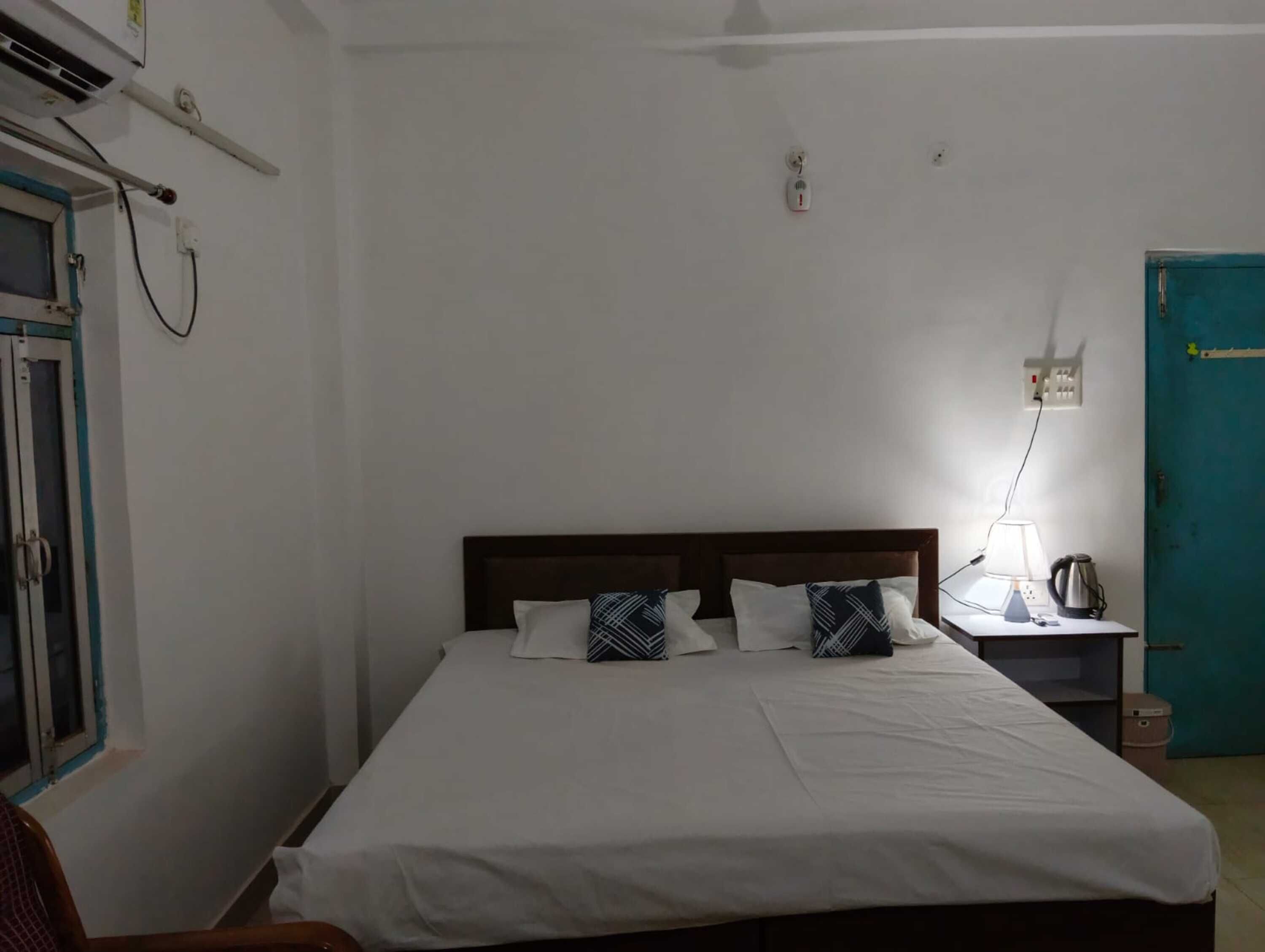 Habitación individual con una cama de madera, ropa de cama blanca, una mesita de noche con lámpara, un hervidor y una puerta azul.