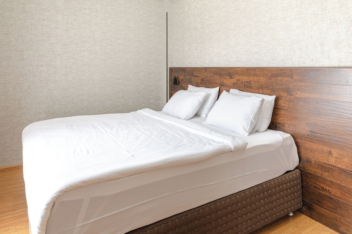 Una cama bien hecha con sábanas blancas, cabecera de madera y pared de color claro.