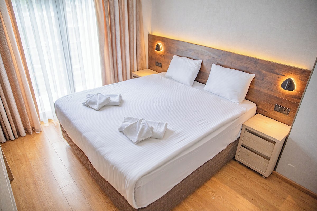 Una cama bien hecha con sábanas blancas y dos almohadas, un cabecero de madera con luces empotradas en la pared y una mesita de noche de madera con un cajón.