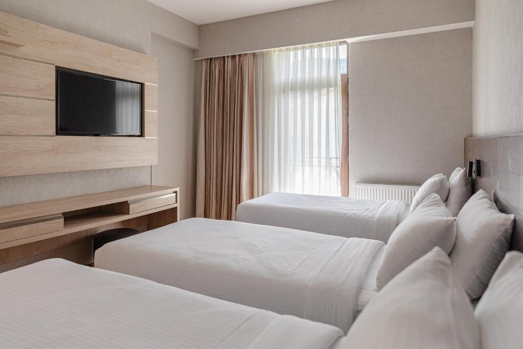 A hotel room with two beds, a flat-screen TV mounted on a wooden panel, and a window with sheer curtains.