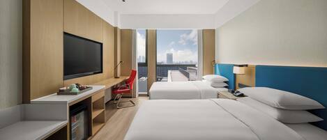 A hotel room with a large TV, two beds, a desk, a red chair, and a city view.