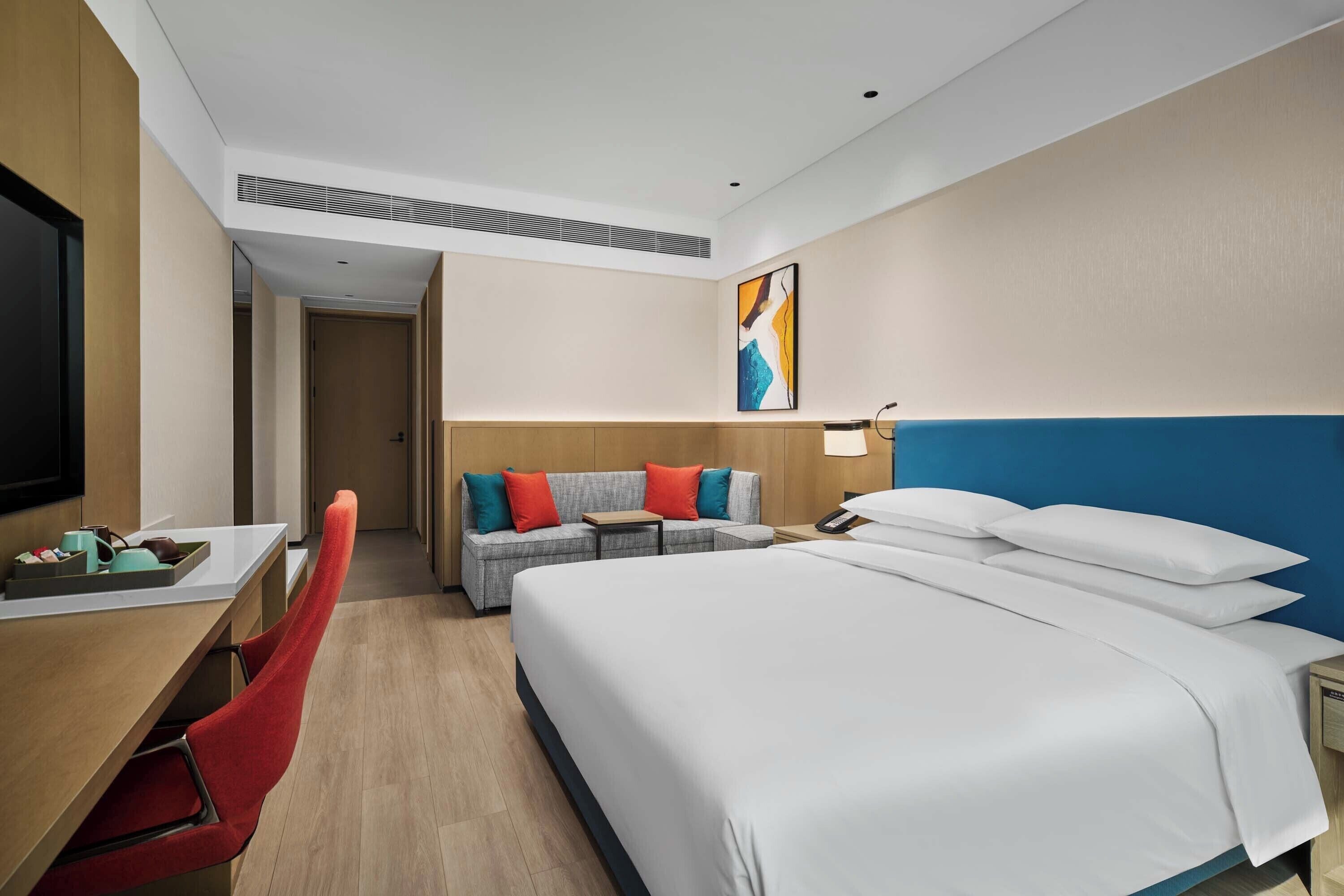 Quarto de hotel com cama, escrivaninha, cadeira, sofá e televisão.