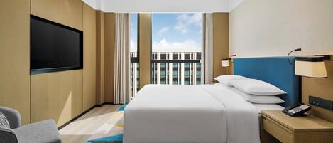 A hotel room with a bed, a TV, a desk with a phone, and a view of buildings through large windows.