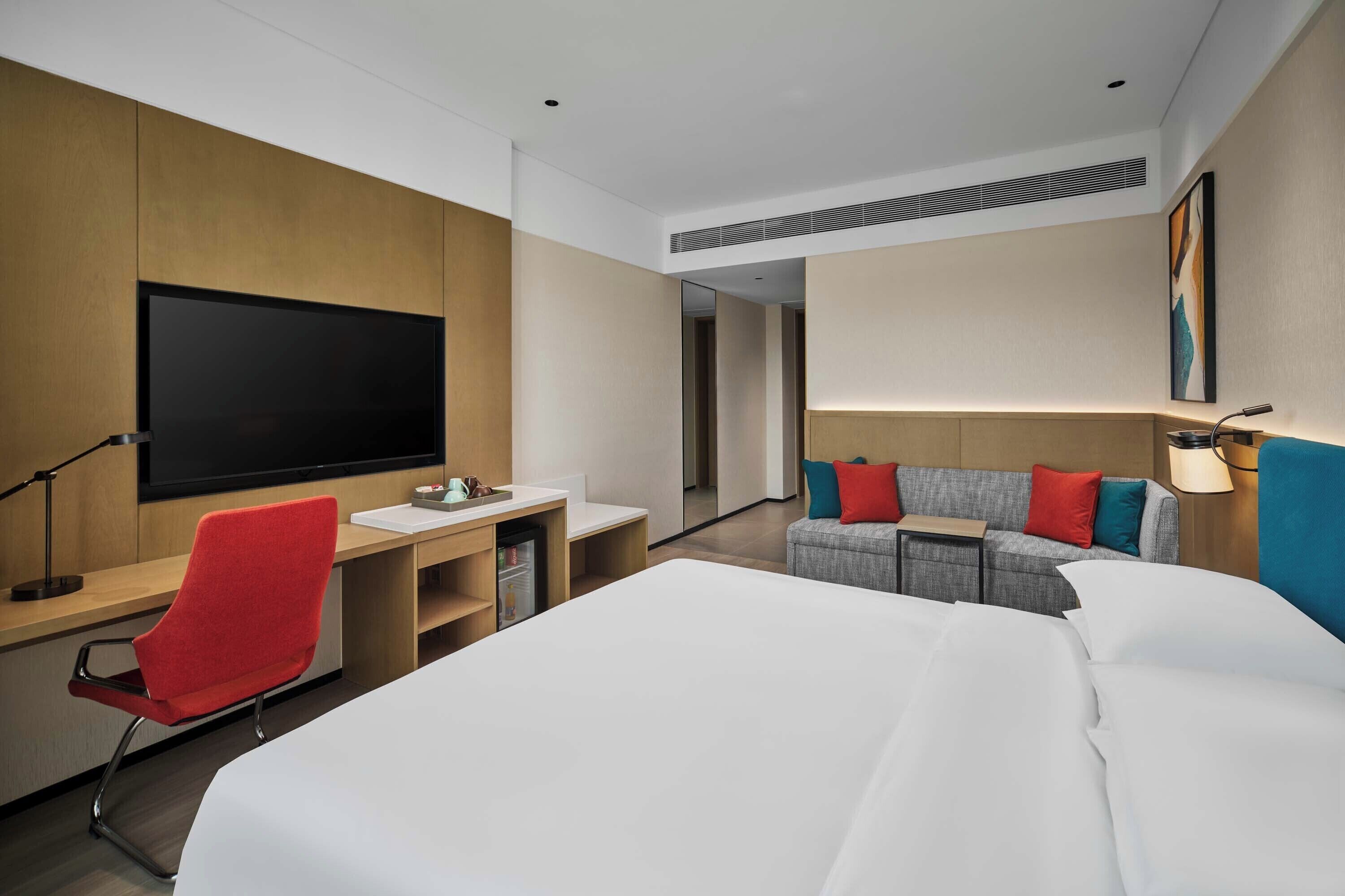 A hotel room with a bed, a desk, a chair, a TV, a sofa, and a lamp.