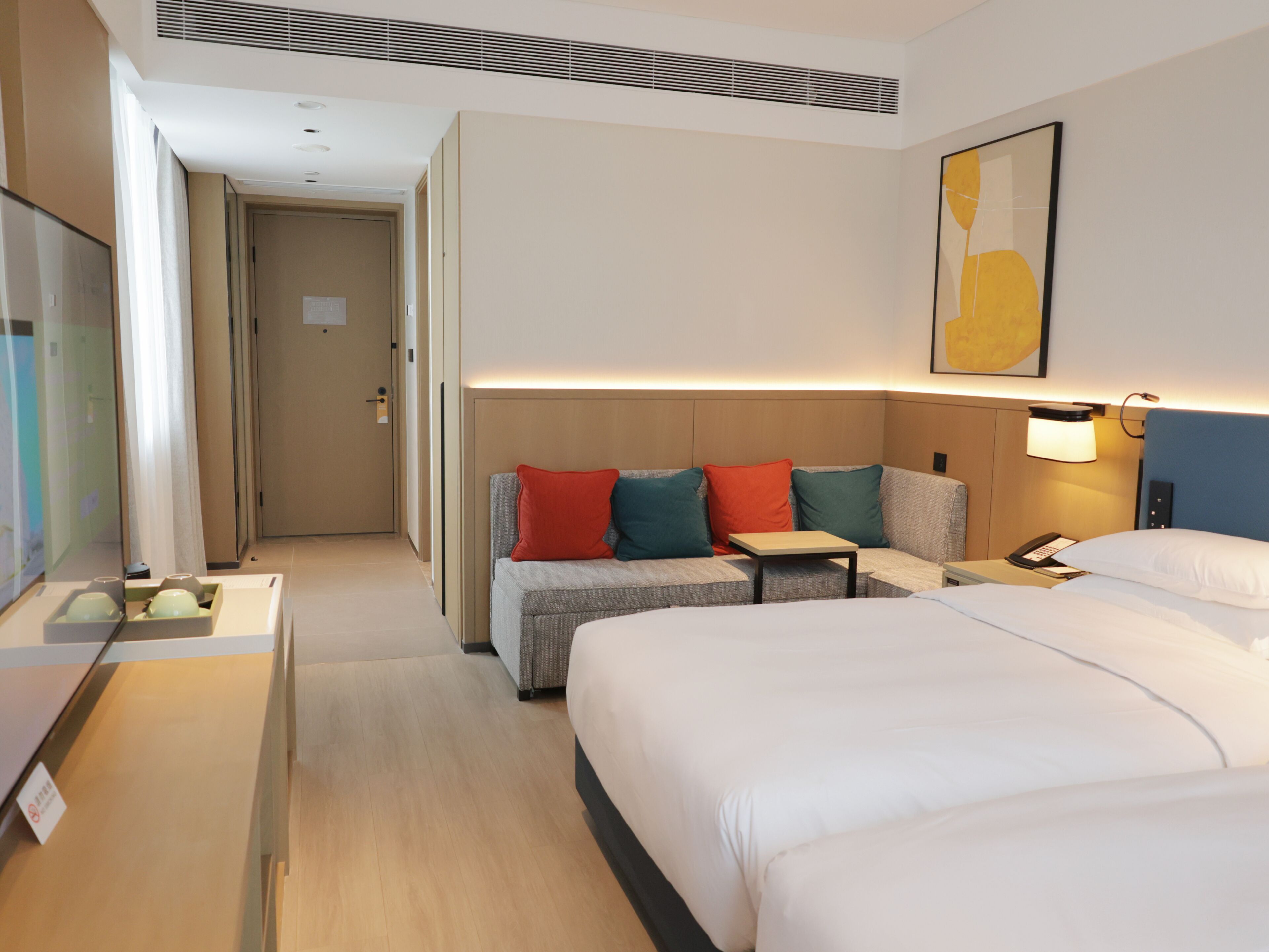 A hotel room with a large bed, a sofa with colorful cushions, a small table, a bedside lamp, and a painting on the wall.