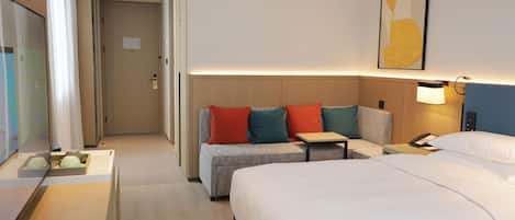 A hotel room with a large bed, a sofa with colorful cushions, a small table, a bedside lamp, and a painting on the wall.
