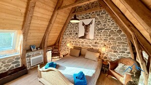 Un dormitorio acogedor en un ático con pared de piedra, techo de madera, una cama con cabecera, una silla e imagen de un ciervo.