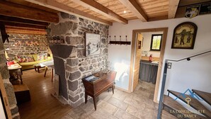 Un interior rústico con pared de piedra, vigas de madera, una zona de estar acogedora y una cocina visible a través de un vano abierto.