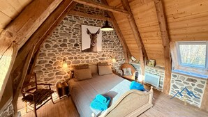 Un dormitorio acogedor en el ático con pared de piedra, cama con toallas azules, chimenea de piedra y ventana con vistas a árboles.