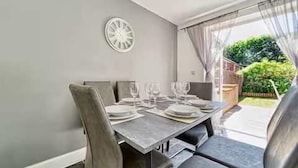 A dining area with a table set for four, a clock on the wall, and a view of a garden through a window.