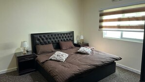 Ein Schlafzimmer mit einem großen Bett, zwei Nachttischen mit Lampen, einem Fenster mit Jalousien und einer Klimaanlage.