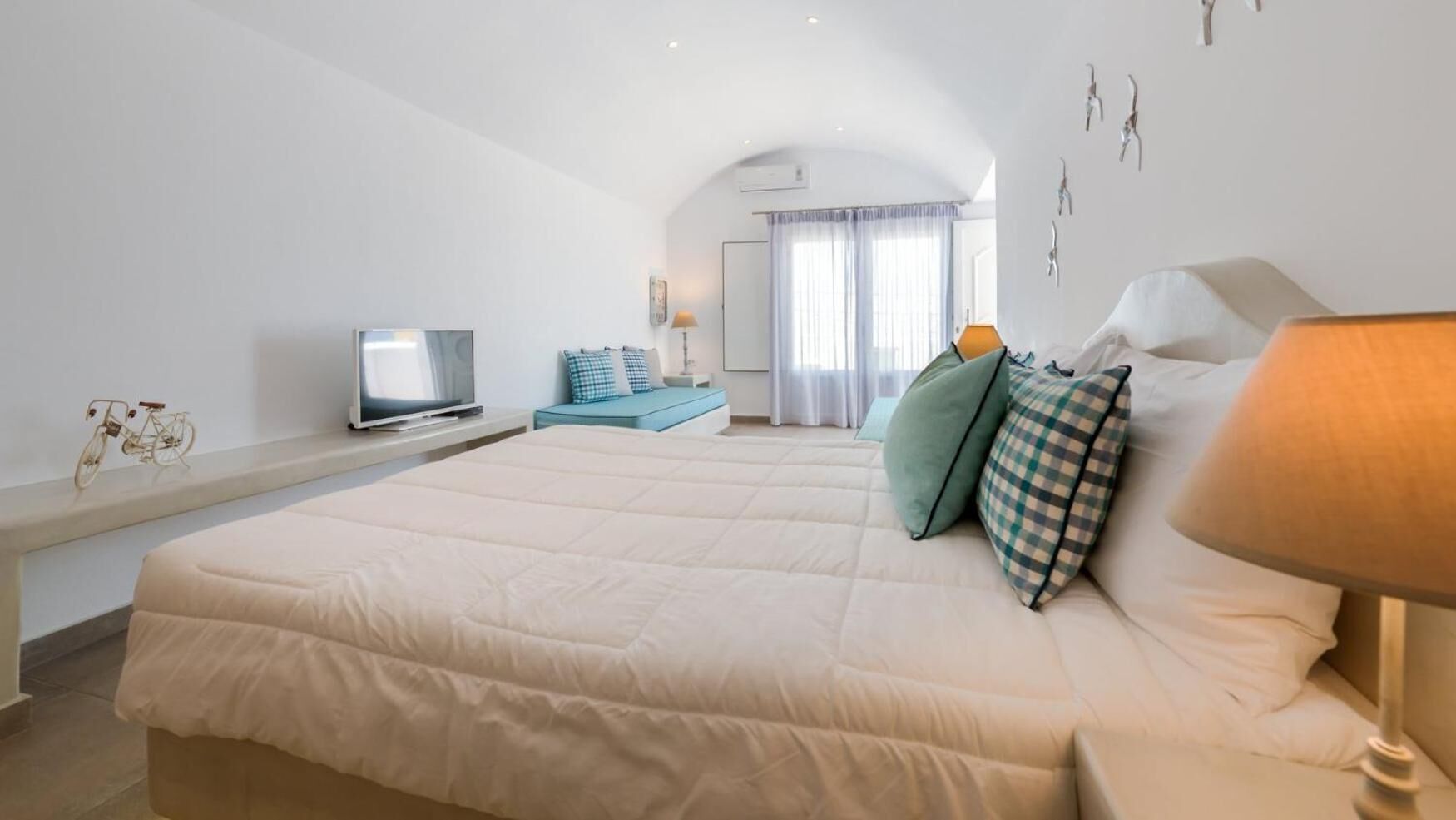 A bedroom with a large bed, a TV on a stand, a desk, and a window with curtains.