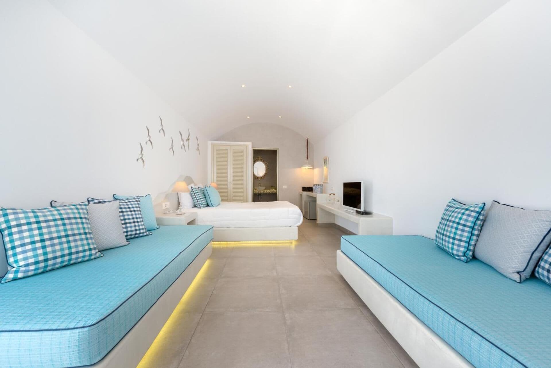 Un dormitorio moderno y bien iluminado con una cama grande, dos sofás, un televisor y un baño visible a través de un arco.